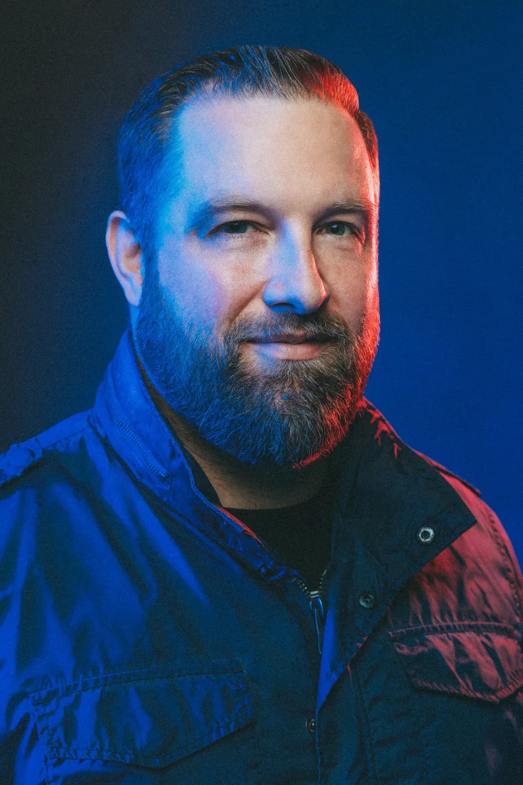 A bearded man in a denim jacket, lit blue and red against a dark backdrop