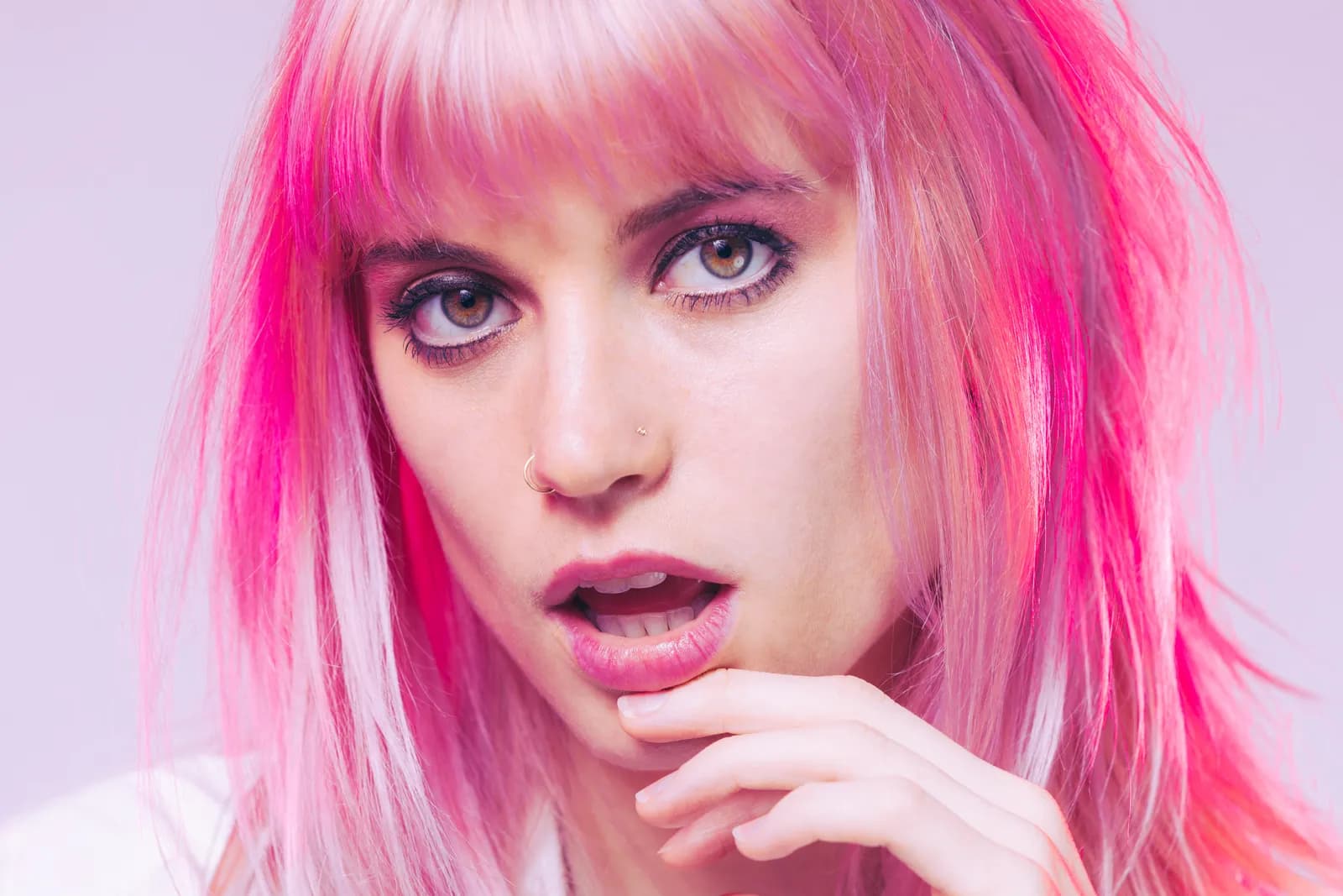 A close-up of a woman with pink hair and a hand at her lip