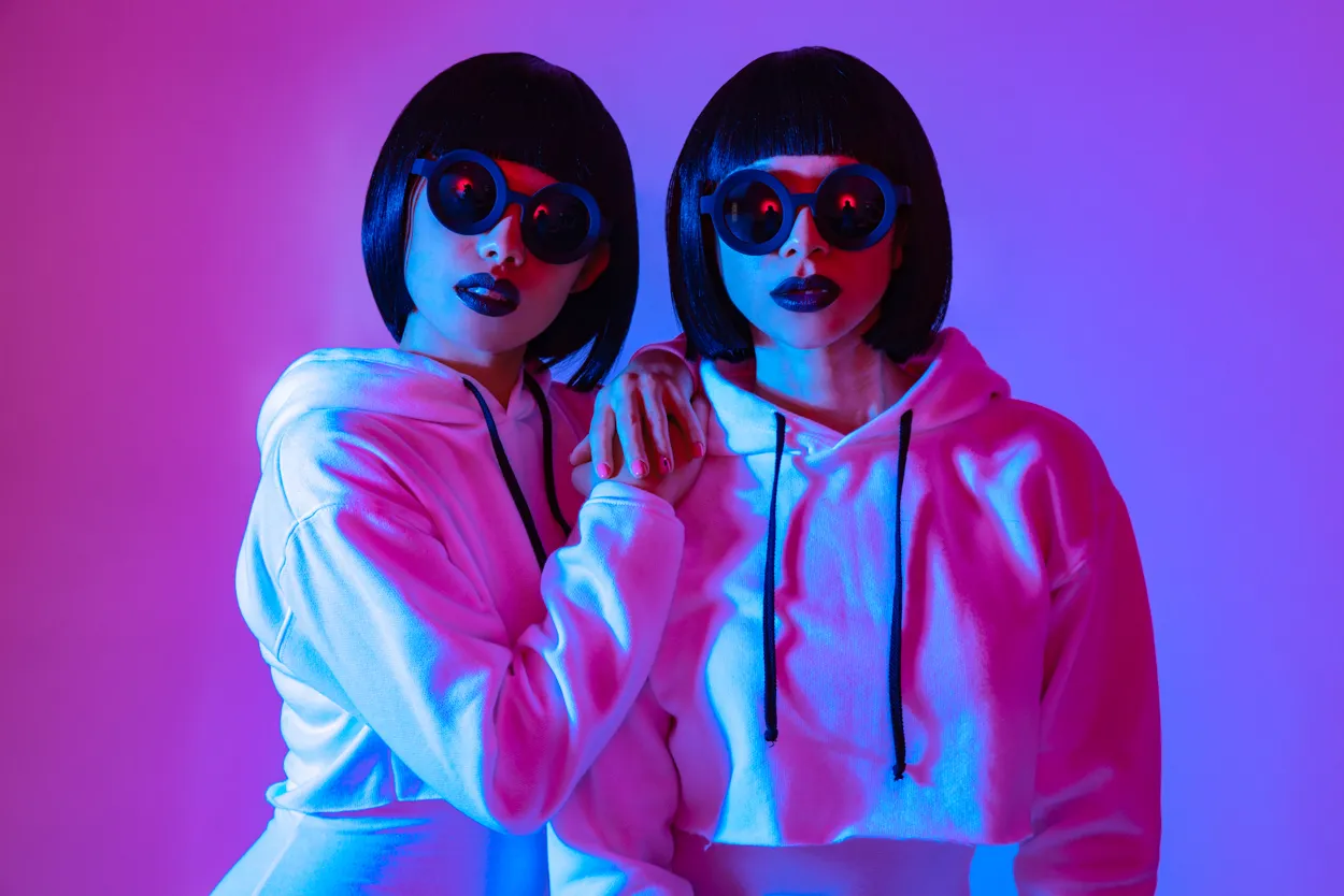 Two women with matching black bobs and sunglasses under pink and blue light