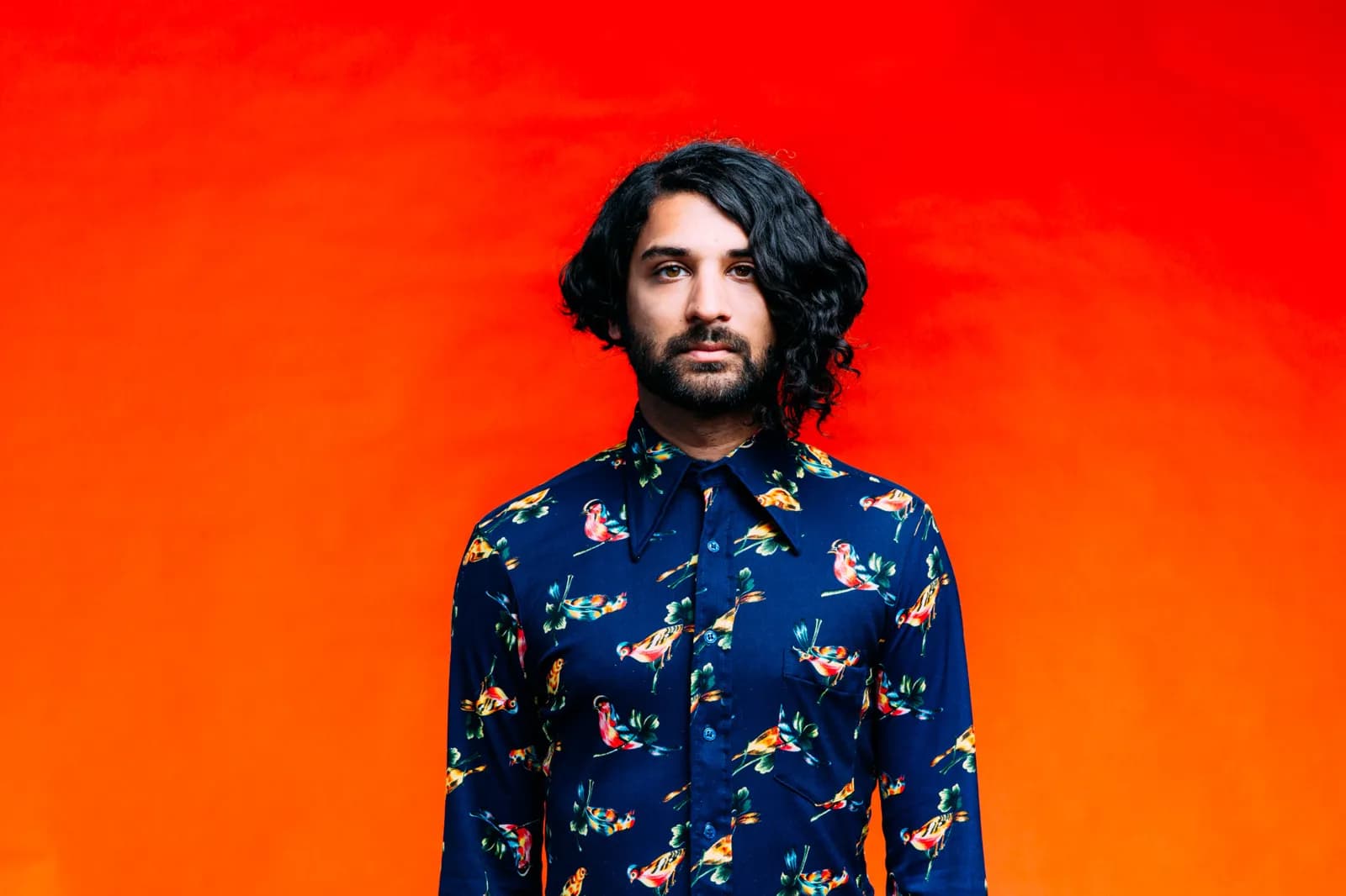 A man with curly hair in a floral shirt against orange and red